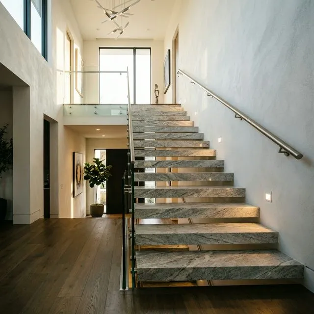 Modern floating stone staircase in light grey granite with glass balustrade and under-tread lighting in a contemporary double-height hallway